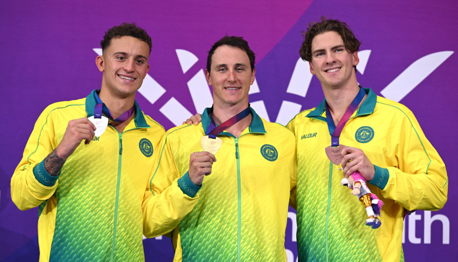 Three male athletes in matching yellow-green jackets pose with their medals against a purple backdrop (left silver, center gold, right bronze).