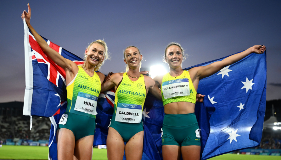 Three Australian female athletes celebrate on the track, holding the Australian flag and a blue flag, smiling under stadium lights.