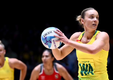 Female volleyball player in a yellow uniform holding a volleyball, ready to pass, with blurred opponents in the background on a dark court.