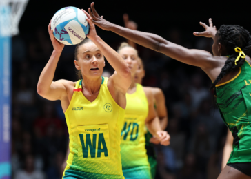 Female volleyball player in yellow jersey holding a ball above her head, preparing to set or spike, on an indoor court as a defender reaches in green near her side.