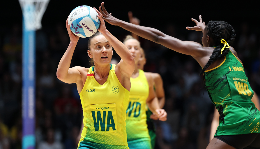 Female volleyball player in yellow jersey holding a ball above her head, preparing to set or spike, on an indoor court as a defender reaches in green near her side.