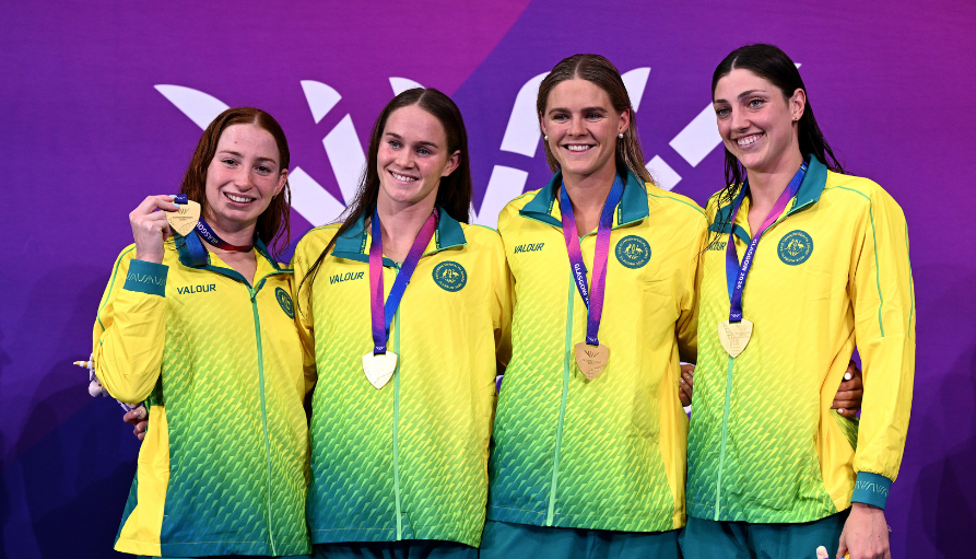 Four female athletes in yellow-green jackets pose with gold medals on a purple backdrop.