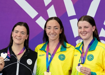 Three female athletes stand on a podium, each displaying a medal; center and right wear yellow jackets with green collars, left wears a black-and-white jacket, all smiling against a purple backdrop.
