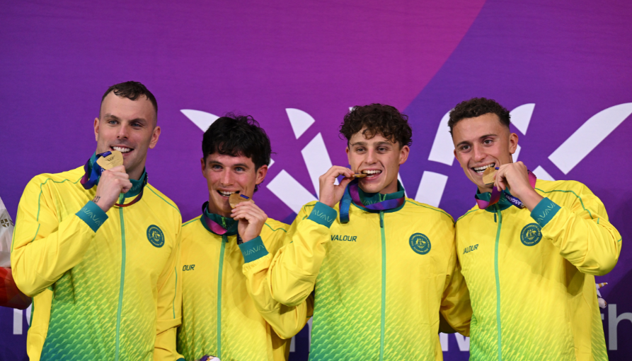 Four male athletes in yellow-green jackets biting gold medals against a purple backdrop.
