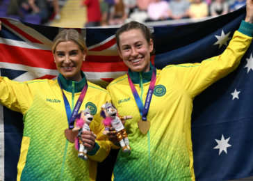Two female athletes in yellow-green uniforms celebrate a win, holding Australian flags and bronze medals with plush mascots on a podium.
