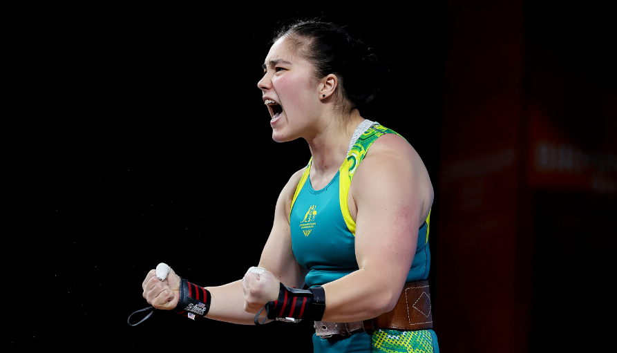 Female weightlifter in a teal singlet wearing a brown weightlifting belt, shouting with clenched fists during competition.