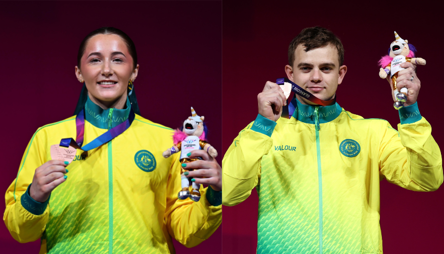Two athletes in neon yellow-green jackets hold medals and a pink-and-white mascot plush on a red stage behind them.
