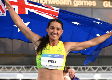 Smiling female athlete in a yellow top raises an Australian flag in celebration at the Commonwealth Games.