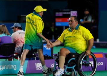 Two male Australian athletes on a sports court share a handshake after a game; one in a wheelchair wearing a yellow-green jersey, the other standing in matching gear, with spectators in the background.
