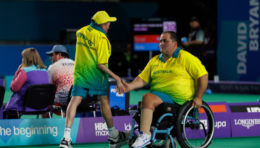 Two male Australian athletes on a sports court share a handshake after a game; one in a wheelchair wearing a yellow-green jersey, the other standing in matching gear, with spectators in the background.