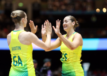 Two female athletes in bright yellow-green uniforms share a high-five on a track, with a cheering crowd in the background.