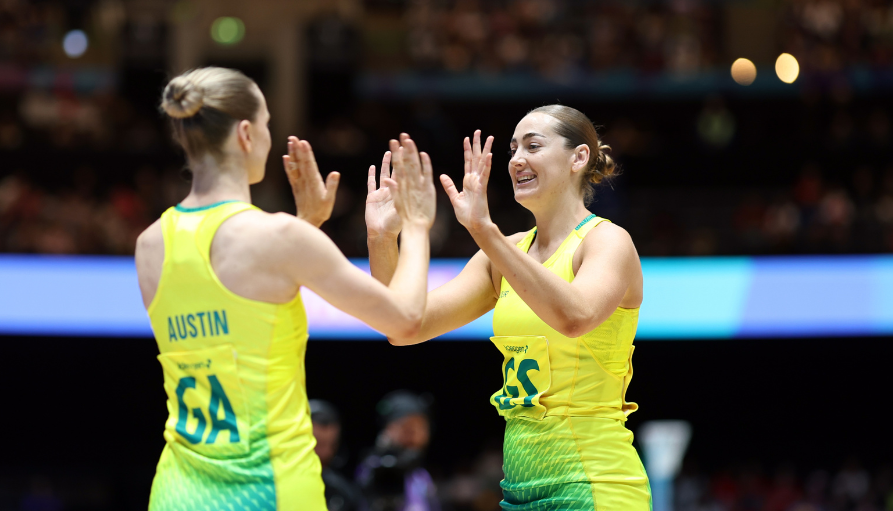 Two female athletes in bright yellow-green uniforms share a high-five on a track, with a cheering crowd in the background.