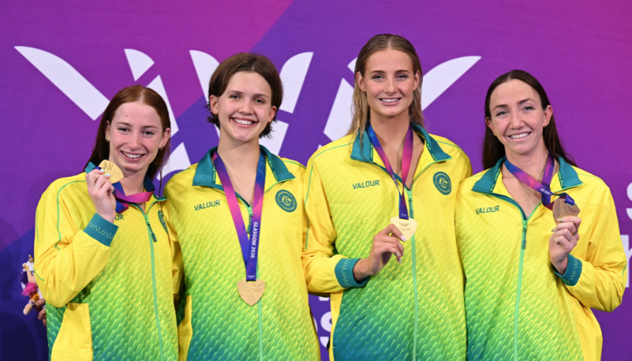 Four female athletes in bright yellow‑green jackets pose with gold medals on purple ribbons against a purple backdrop; smiles proudly after a podium finish.